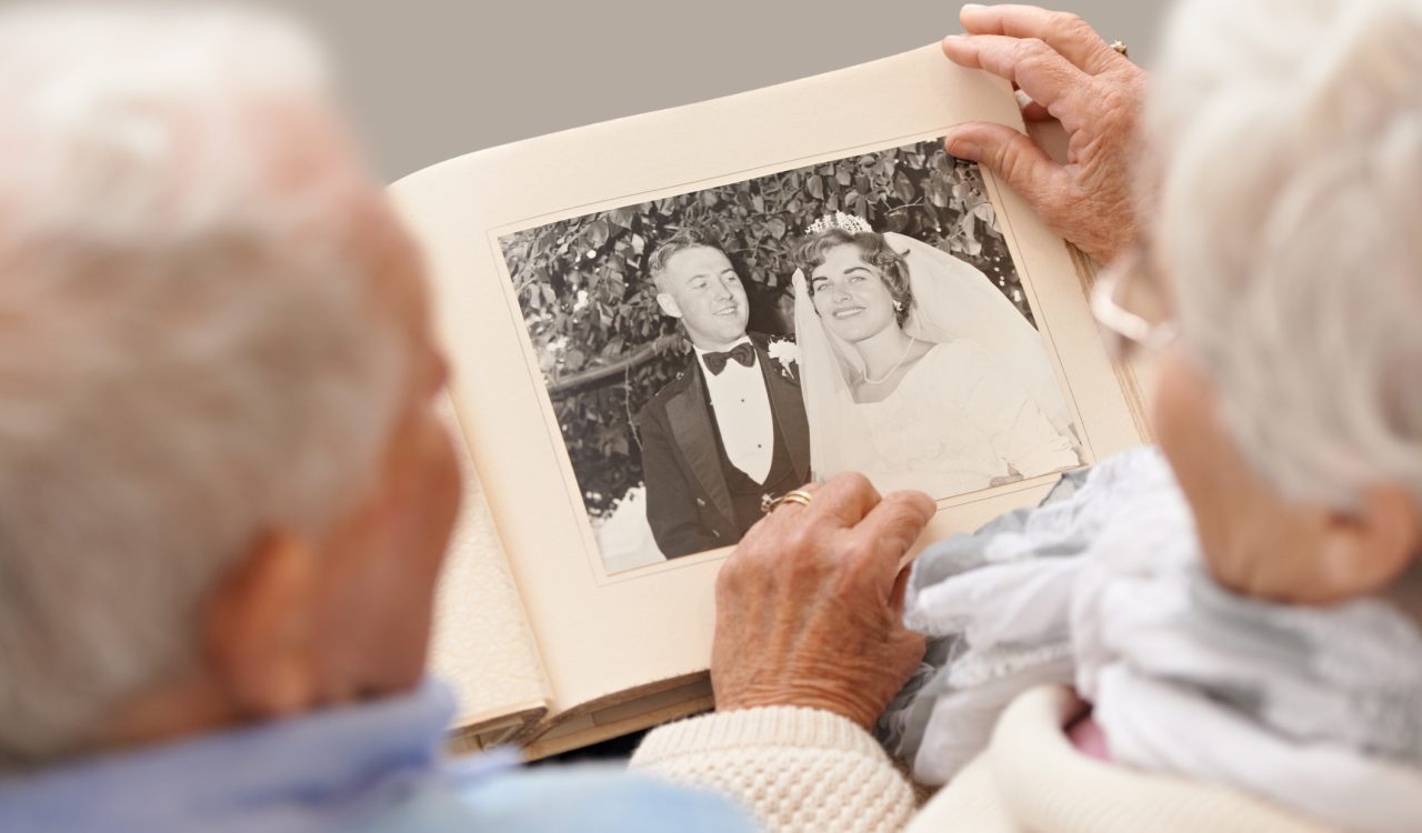 Blurred silhouettes of the back of an older adult man and older adult woman’s heads as they sit down holding an album with their wedding photo taken when they were young.