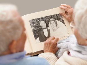 Blurred silhouettes of the back of an older adult man and older adult woman’s heads as they sit down holding an album with their wedding photo taken when they were young.