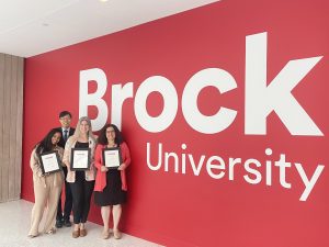 A group of people stand in front of a red wall with the words "Brock University" painted on it.