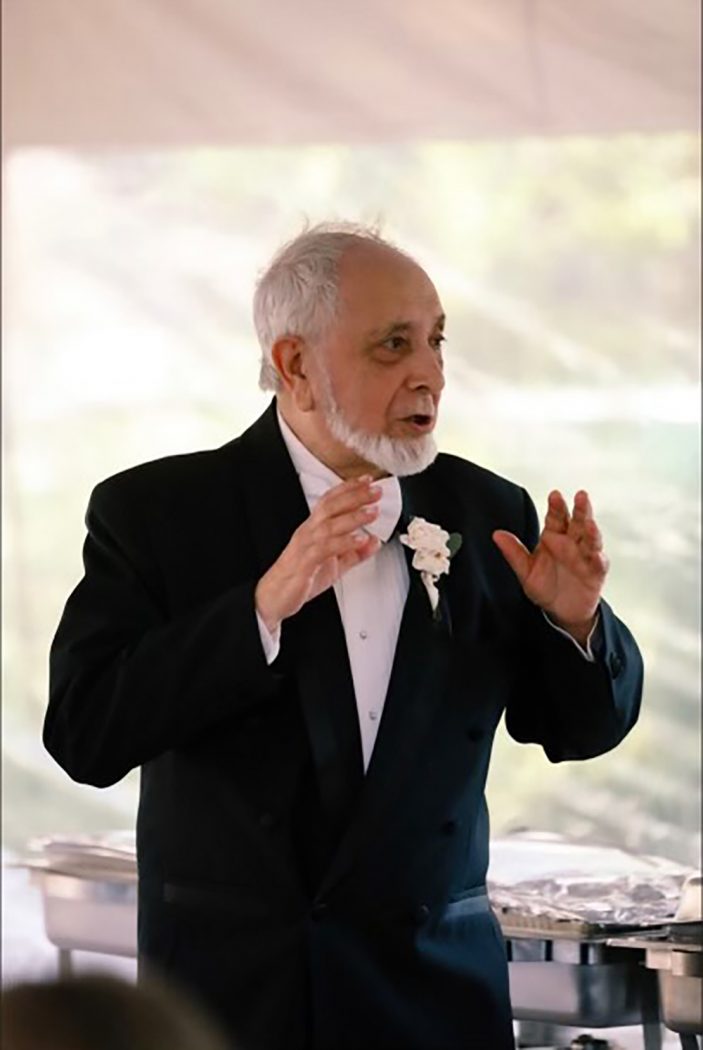 Juan Fernandez as an older gentlemen in a black tuxedo with a white floral corsage. He is mid-speech with his arms raised in the air with a kind face set against a white backdrop.