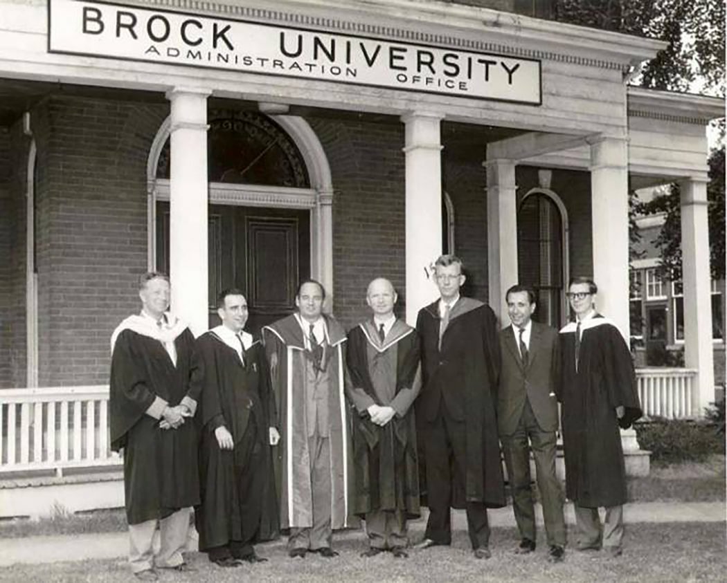 A black and white archival photo shows seven Brock University professors dressed in regalia standing shoulder to shoulder in front of a sign that reads “Brock University Administration Office”. The seven men are all smiling standing outside of a brick building with a white fence.