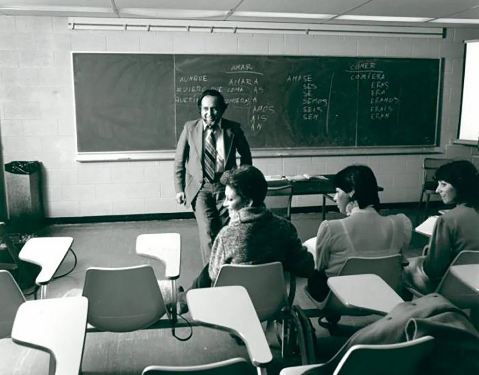 In a black and white photo of a university classroom, Brock Spanish professor Juan Amadeo Fernandez leans against a desk smiling at three students sitting in front of him.