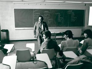 In a black and white photo of a university classroom, Brock Spanish professor Juan Amadeo Fernandez leans against a desk smiling at three students sitting in front of him.