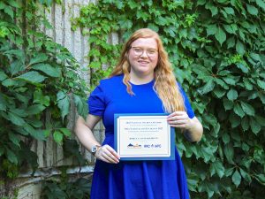 A woman in a blue dress stands in front of a leafy background while holding a certificate.