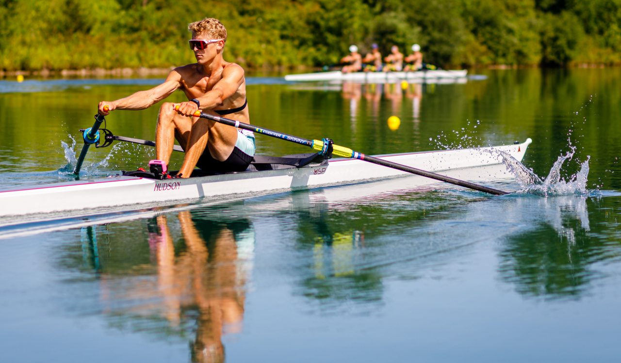 An athlete rows a boat on water backdropped by green trees.