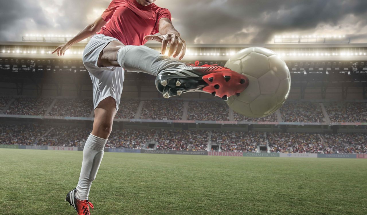 A close up image of woman soccer player kicking ball in a outdoor floodlit stadium full of spectators under a stormy evening sky.