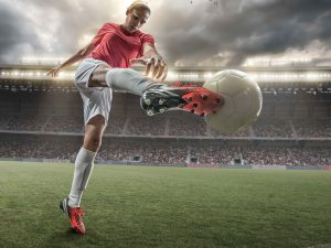 A close up image of woman soccer player kicking ball in a outdoor floodlit stadium full of spectators under a stormy evening sky.