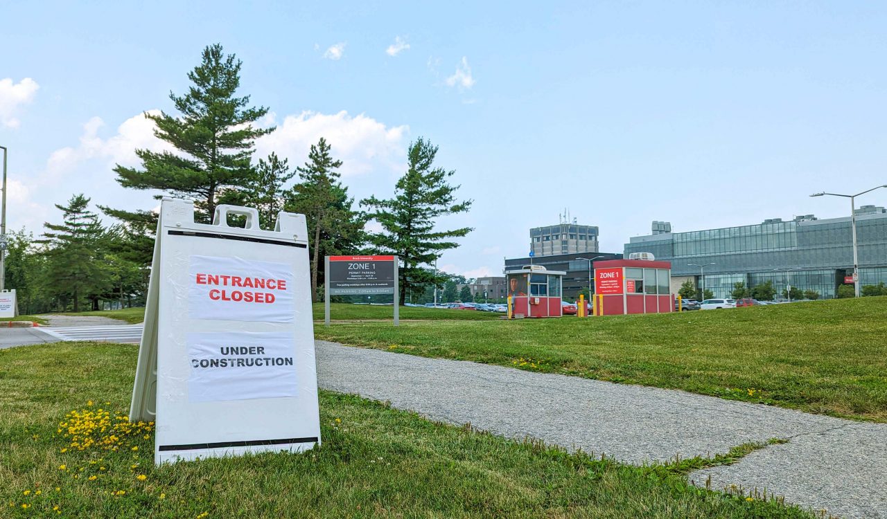 A sign that says ‘Entrance Closed, Under Construction’ sits on a a boulevard of grass in front of a parking lot entrance.