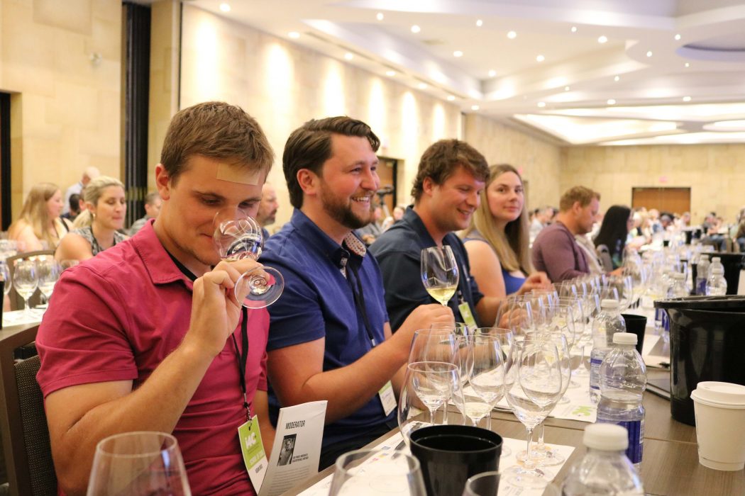 Seated at a table in a conference room, Aidan Buis smells wine in a glass while Benjamin Whitty looks forward holding a wine glass and Keelan Buis and Morgan Buis look on.