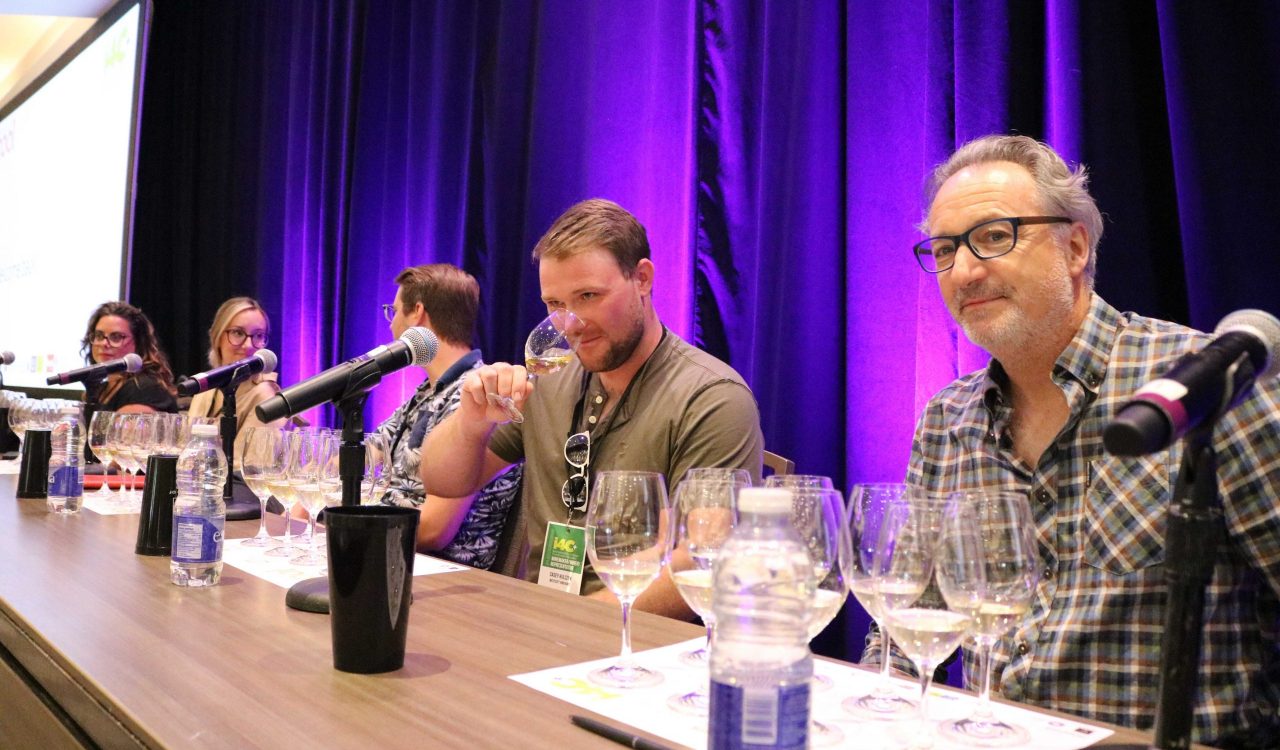 Five people sit at a table on a stage with purple curtains behind them. In front of each person is a microphone and eight wine glasses.