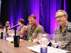 Five people sit at a table on a stage with purple curtains behind them. In front of each person is a microphone and eight wine glasses.