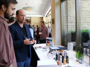 Two men discuss the wine supplies displayed in the table in front of them including a row of bottles, corks and marketing materials.