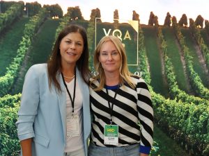Two women pose in front of a banner with an image of rows of grape vines on it.
