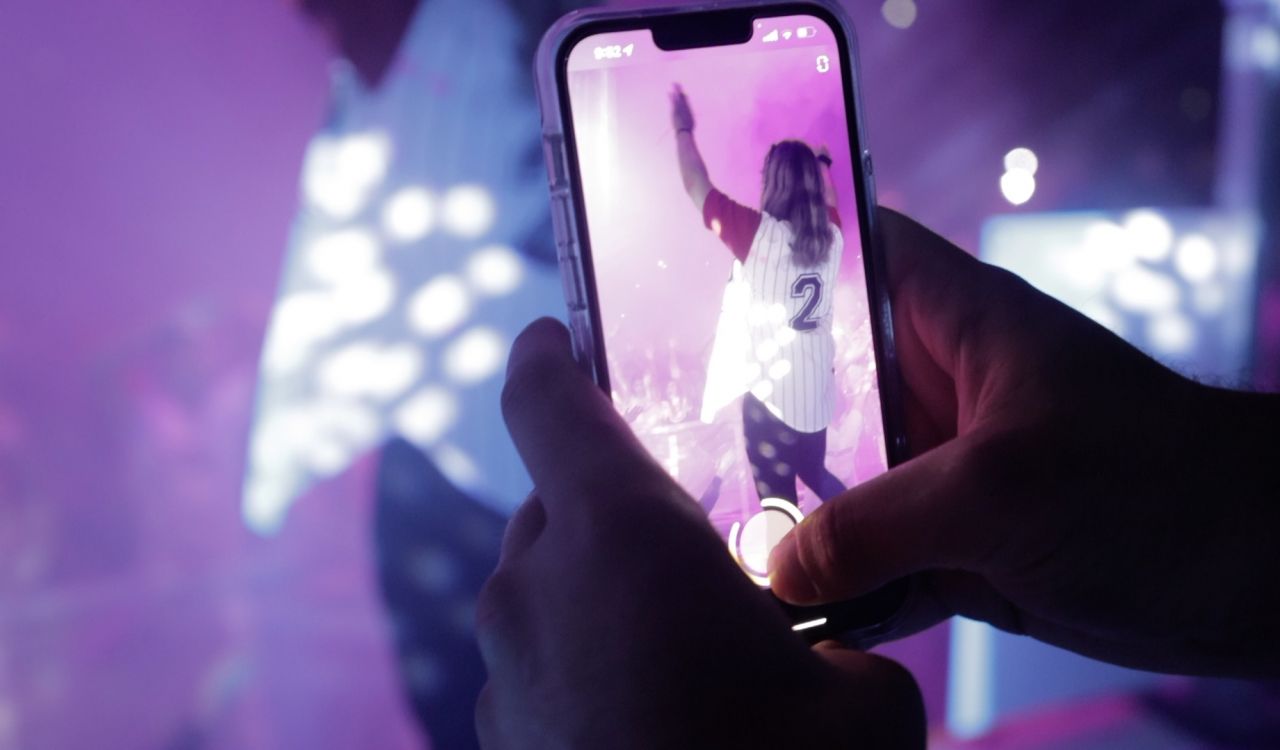 A hand holding a smartphone showing someone performing on stage through the screen.