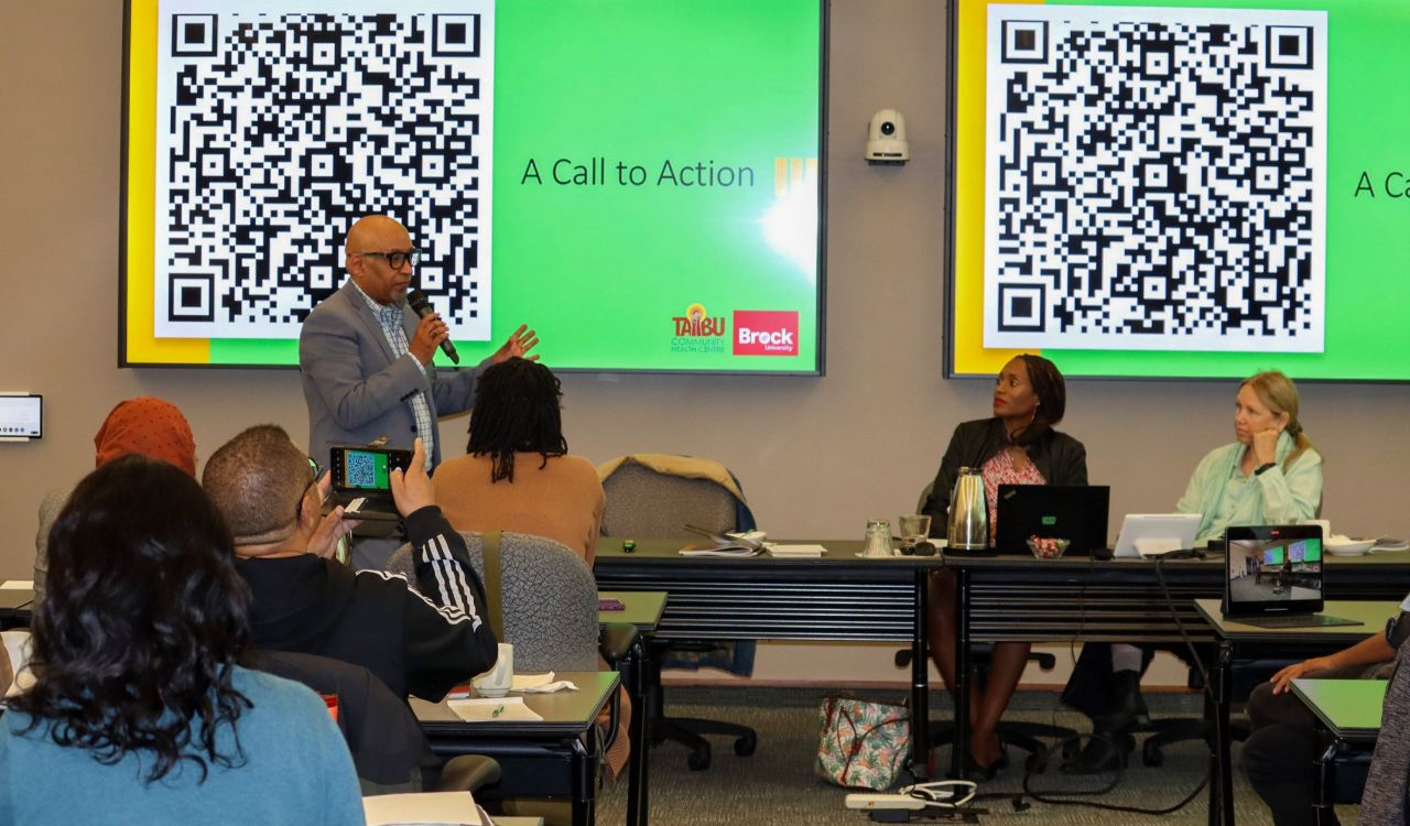 Liben Gebremikael stands with a microphone in front of a green screen with a large QR code. Beside him, seated, are Sadie Goddard-Durant and Andrea Doucet in front of a second screen with the same image.