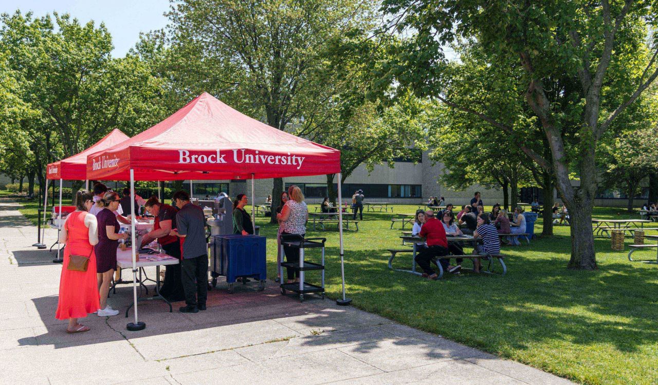 People gather at picnic tables and collect food from under a red awning in a courtyard.