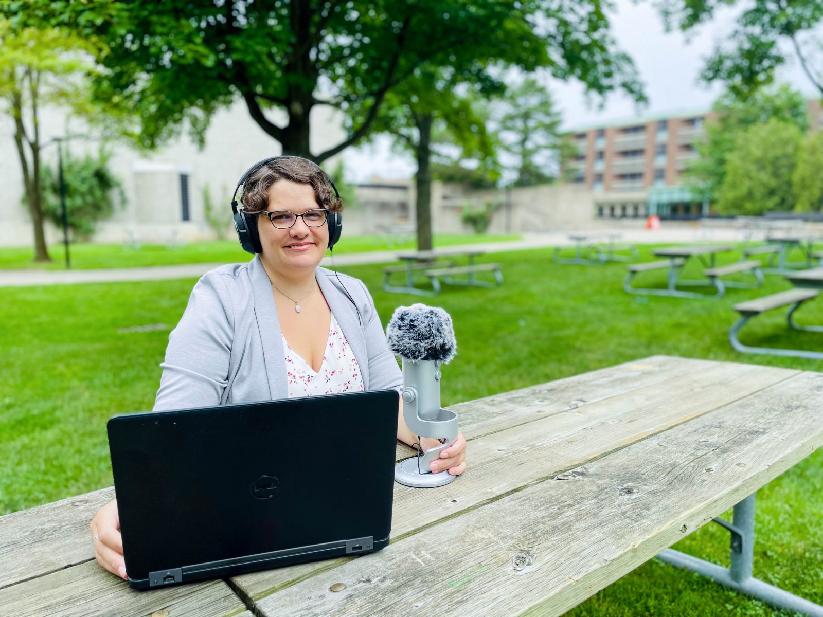 A woman with short hair and wearing glasses smiles warmly at the camera while sitting outdoors at Brock University in Jubilee Court. She is wearing a grey blazer and is sitting at a picnic bench, with a black laptop, a cell phone and recording equipment and microphone sitting in front of her. Green grass and empty picnic tables can be seen behind her.