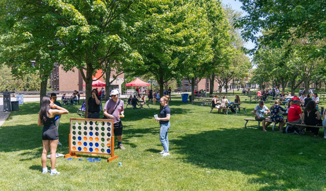 A group of people stand playing lawn games while surrounded by other people sitting at picnic tables in an outdoor courtyard.