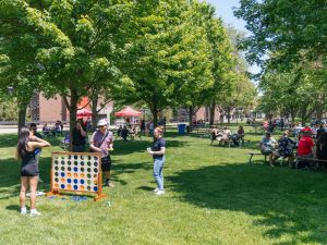 A group of people stand playing lawn games while surrounded by other people sitting at picnic tables in an outdoor courtyard.