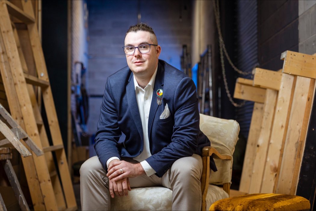 Professional portrait of Deneh’Cho Thompson (he/they) sitting backstage in a theatre. A beaded medicine wheel pin is on the lapel of their navy suit jacket.