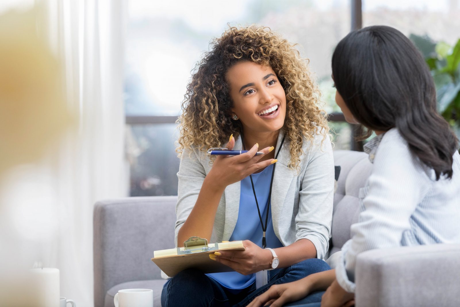 A young female therapist gestures as she talks with a female client. The therapist smiles warmly as she talks with the young woman.