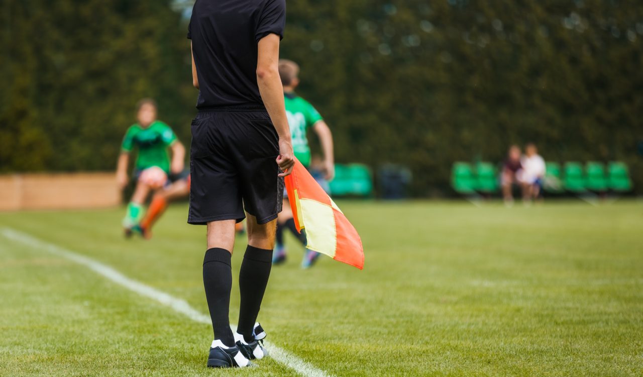 A youth soccer referee stands on the sidelines of a junior-level soccer match while holding a flag.