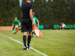 A youth soccer referee stands on the sidelines of a junior-level soccer match while holding a flag.