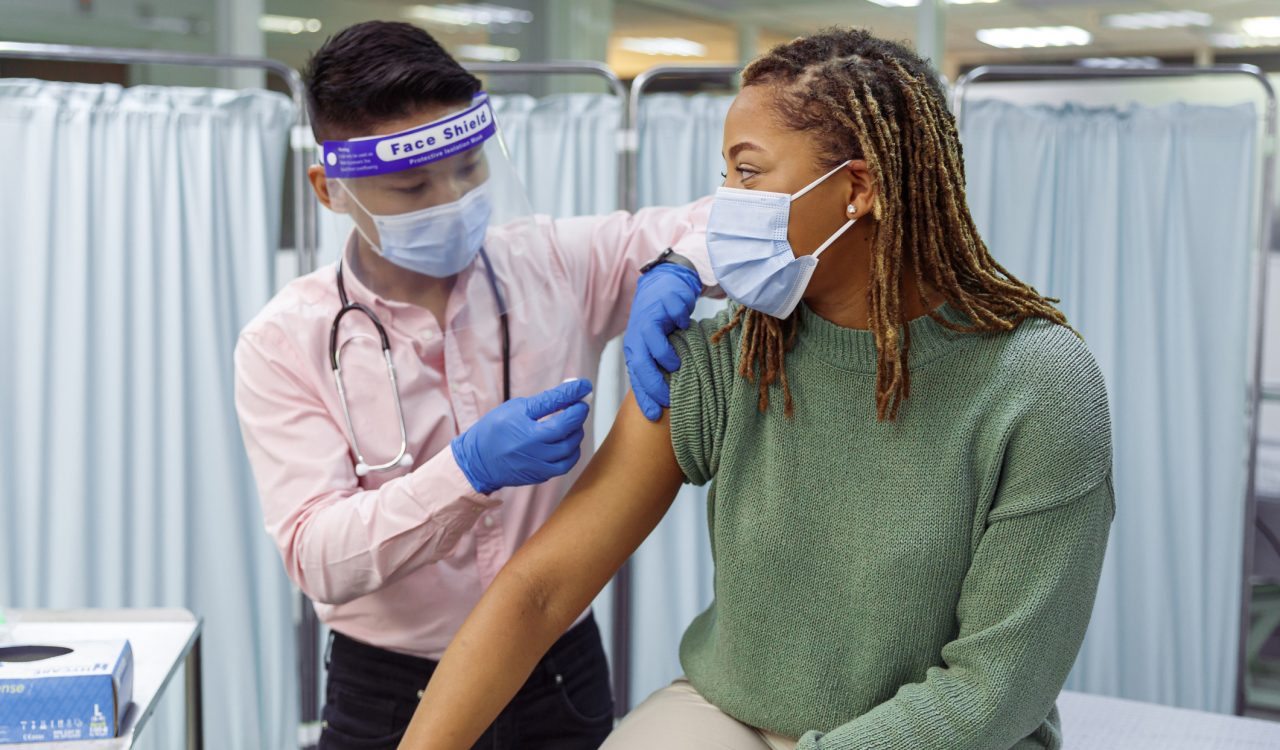 A young Black woman wearing a protective face mask sits in a medical clinic as a doctor prepares to give her an injection in the upper arm.