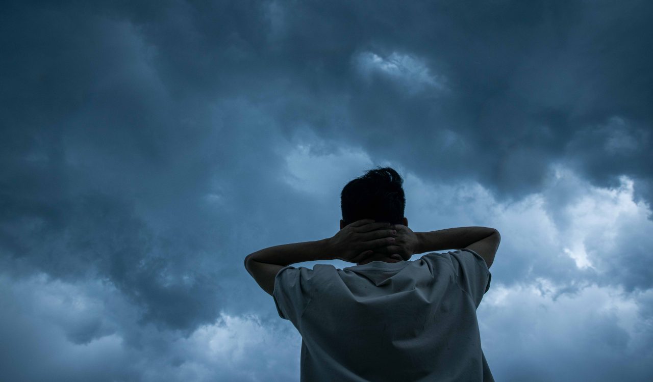 A man holds his hands against his head while looking up at a stormy sky.