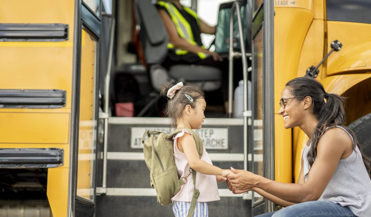 A woman bends down and holds the hands of a nervous little girl as she encourages her before getting on the bus to go home. The little girl is dressed casually and has a backpack on as the driver waits patiently for her to board the bus.
