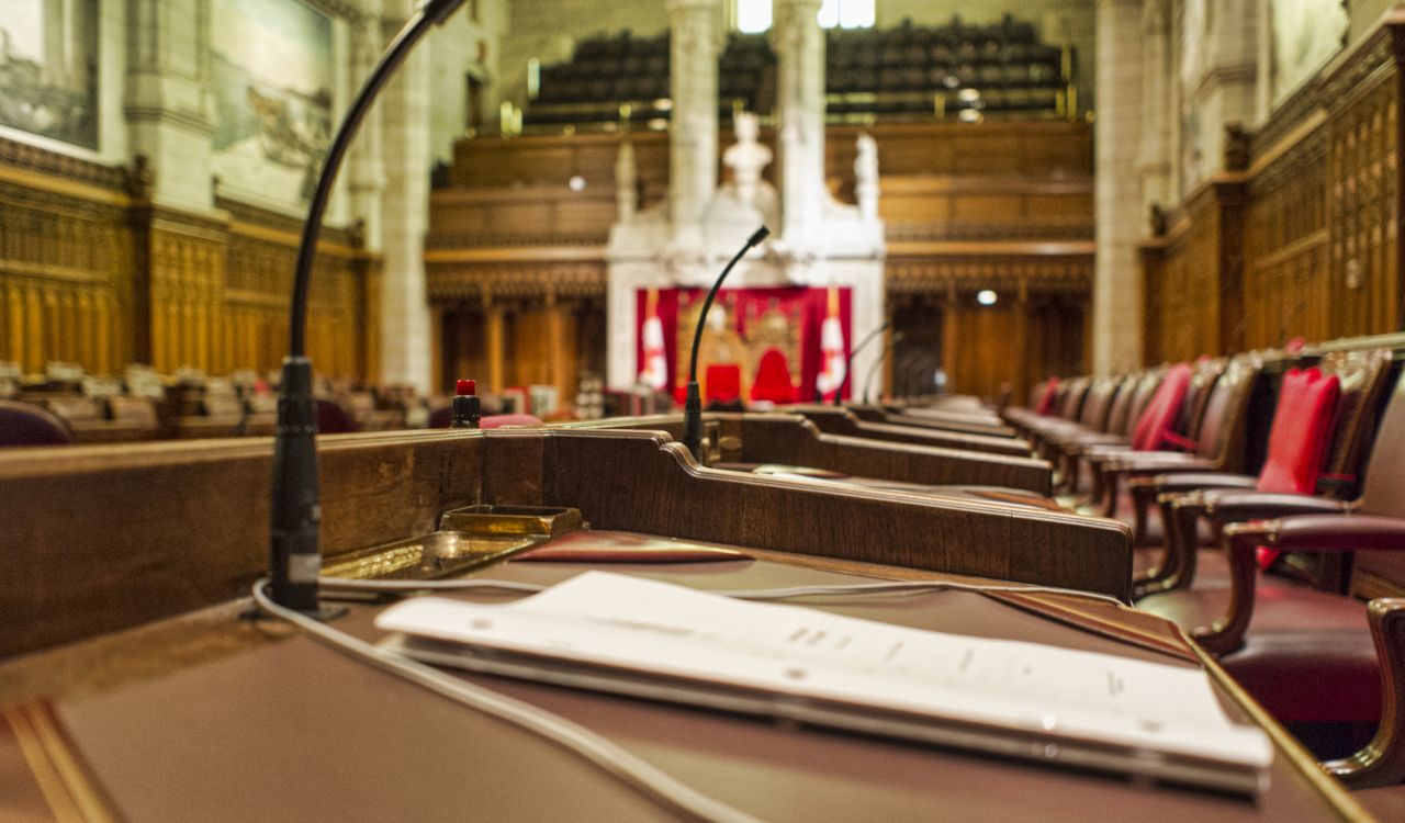 Close up of a Senator's desk with paperwork in the chambers of the Senate of Canada. The Speaker's chair can be seen in the background.