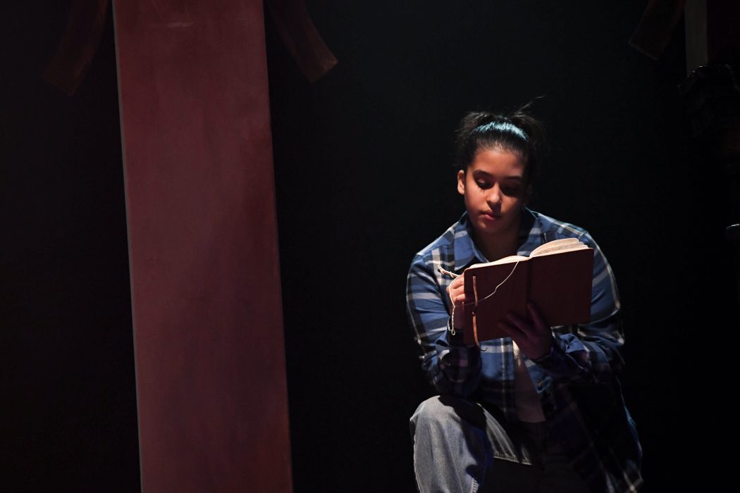 Costumed in a plaid shirt, Actress Hayley King focuses on writing in a notebook. Dramatic theatrical lighting casts part of her face in shadow.