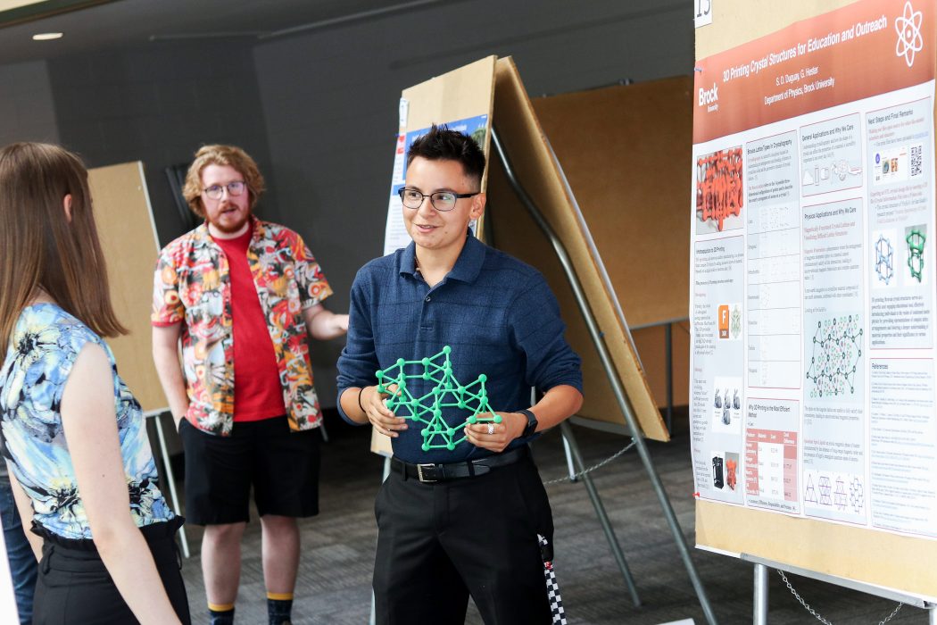 Brock University Physics student Sebastien Duguay holds a 3D printed crystal structure as they speak to a fellow student about their research on “3D Printing Crystal Structures for Education and Research.” Behind them is a large poster outlining their research. 