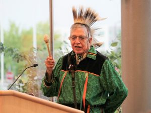 A First Nations man dressed in traditional regalia stands at a podium holding a drumstick addressing delegates at a research event.