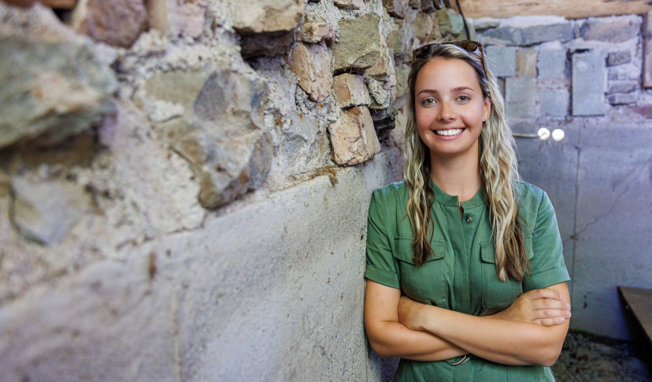 A portrait shows a woman with long blonde hair and a green dress with her arms crossed smiling warmly at the camera while leaning against an old heritage home wall made up of stones in various colours.