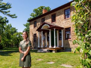 A woman with long blonde hair and a green dress stands outdoors on green grass with a blue sky. Behind her is a The Brown Homestead, a heritage home with red stone, big windows and a grand entranceway to the house.