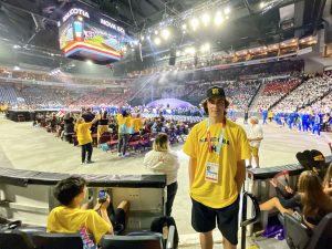 A young man, Lucas Rotondo, wearing a yellow Team Manitoba T-shirt and hat poses for a photo in a sport arena filled with spectators at the North American Indigenous Games in Halifax Nova Scotia.