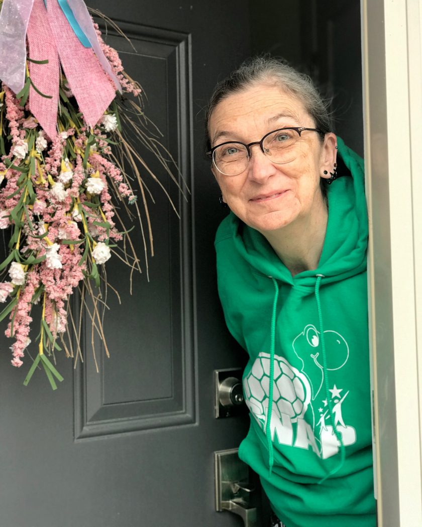 Maureen Connoly peeking out a front door adorned with pink and green florals. Connolly is wearing a green hoodie with an image of a cartoon turtle and the acronym “SNAP.”
