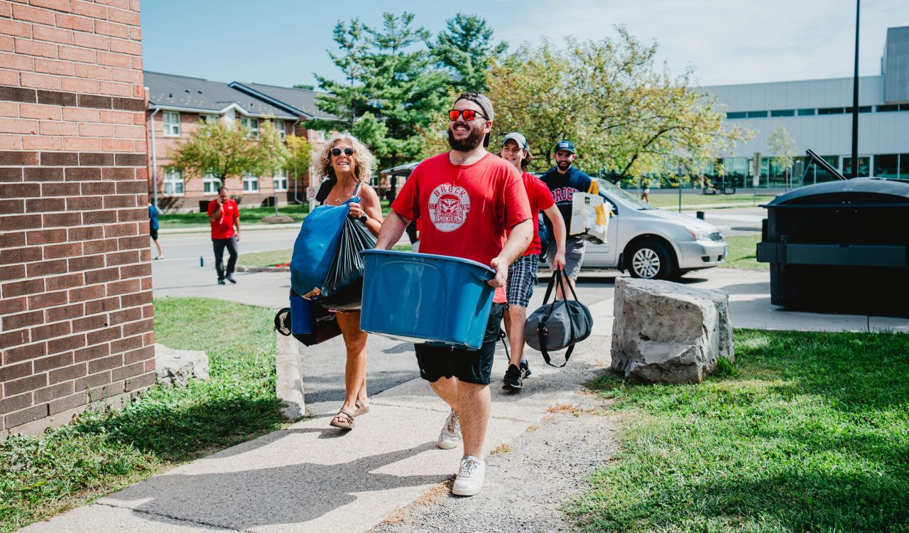 A man carries a large blue tote while others in the background also carry bags while moving into a building.