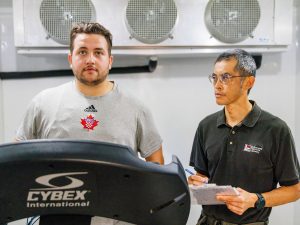 A man on a treadmill is observed by a researcher holding a clipboard.