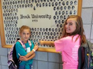 A collage of several Brock graduation photos hangs on a wall. Two young girls point to a photo of a man.