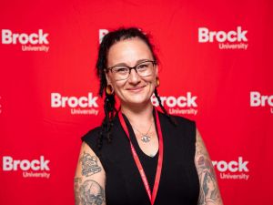 Brock alumna Mahoganie Hines stands in front of a red wall decorated with Brock University logos.