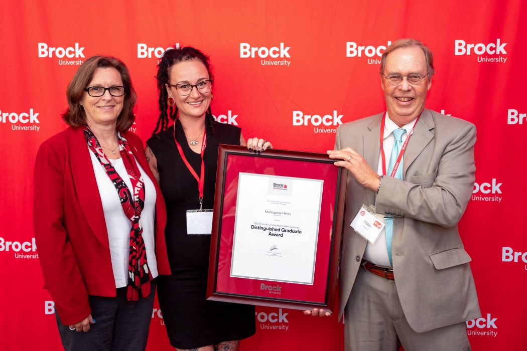 Three people stand side-by-side in front of a red wall decorated with Brock University logos. 