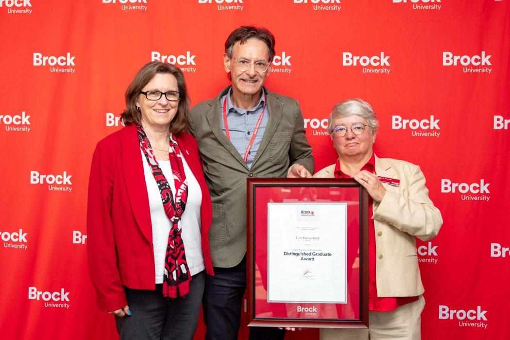 Three people pose in front of a red wall with a framed award certificate. 