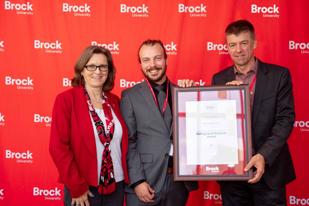 Three people pose side-by-side in front of a red wall. Two of them are holding a framed certificate between them. 