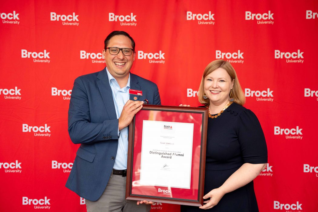 A man and a woman pose holding a framed certificate in front of a red wall. 