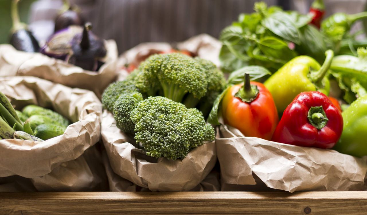 Fresh broccoli, peppers and other vegetables are displayed in paper bags on a wooden table.