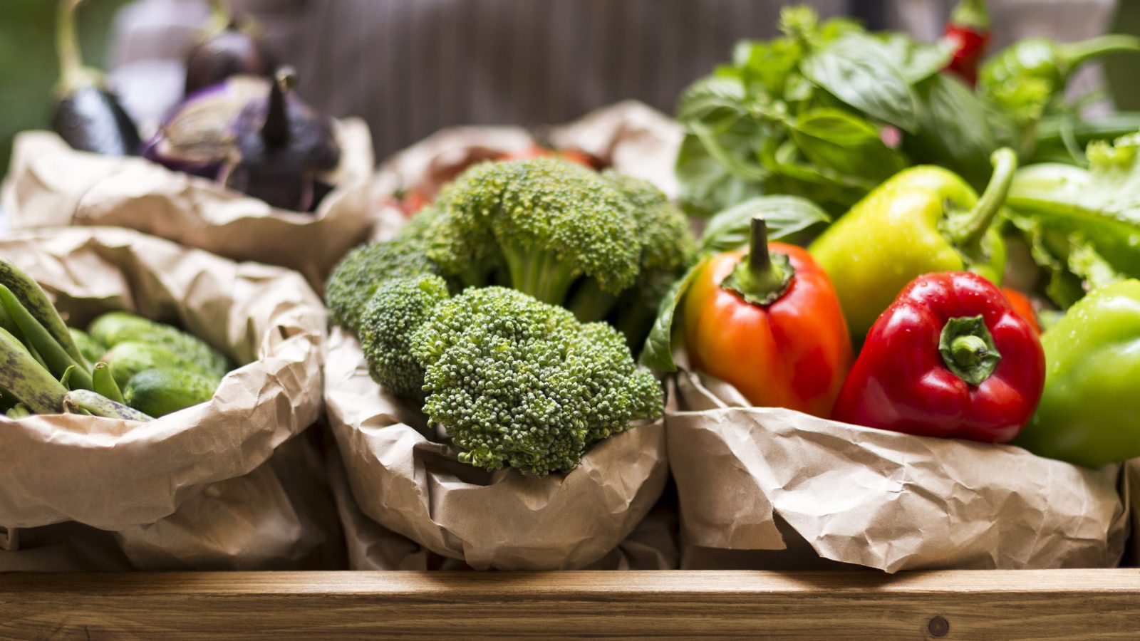 Fresh broccoli, peppers and other vegetables are displayed in paper bags on a wooden table.