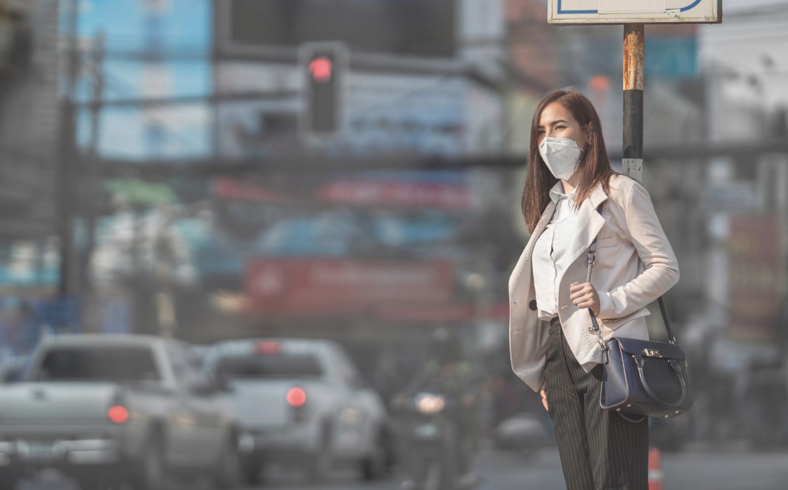 A woman stands at a bus stop, clutching her purse and wearing an N95 mask, with a blurred intersection, vehicles, traffic lights and buildings in the background.