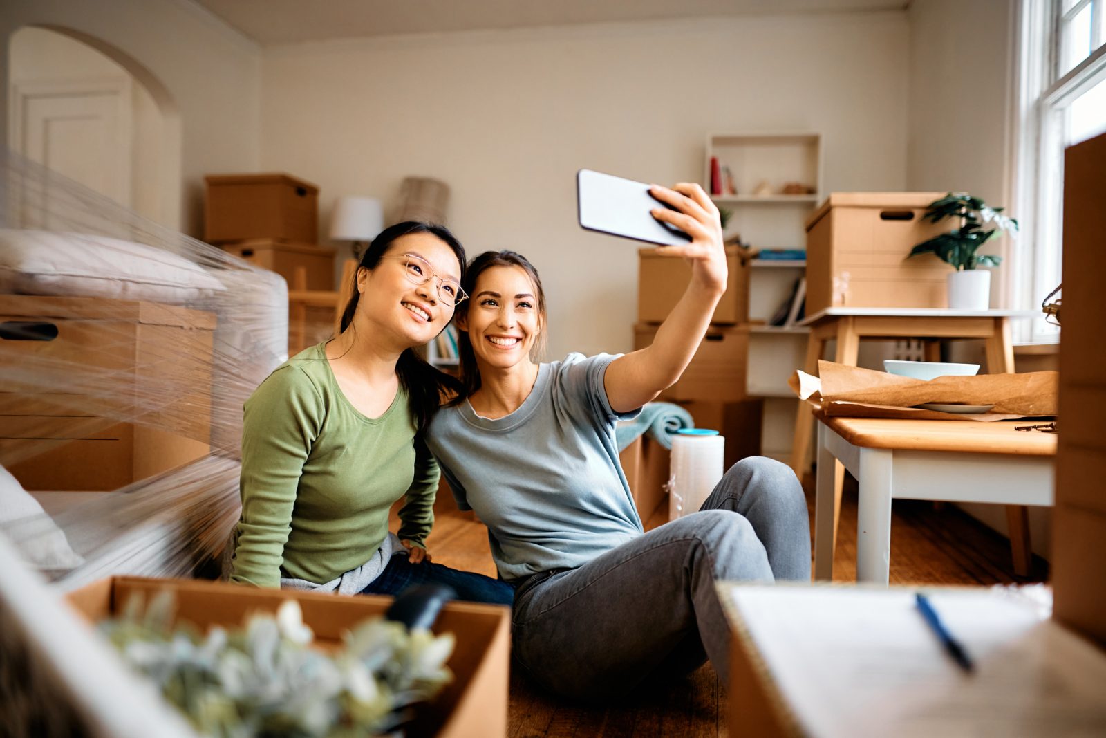 Two young women take a selfie while sitting in the middle of a room full of cardboard moving boxes.
